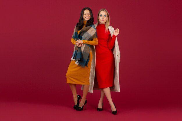 Two stylish women in autumn winter fashion dress and coat posing isolated  on red wall 