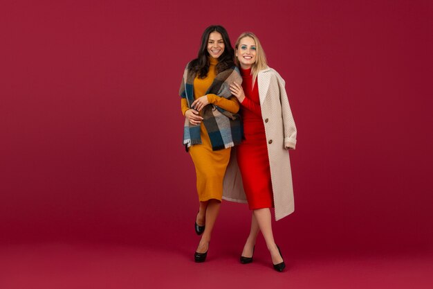 Two stylish women in autumn winter fashion dress and coat posing isolated  on red wall 