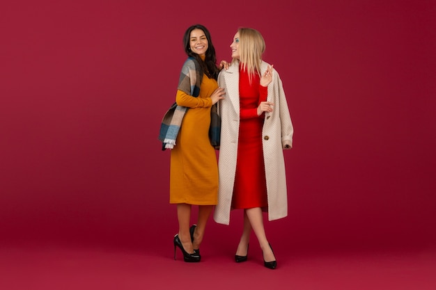 Две стильные женщины в осенне-зимнем модном платье и пальто позирует изолированы на красной стене