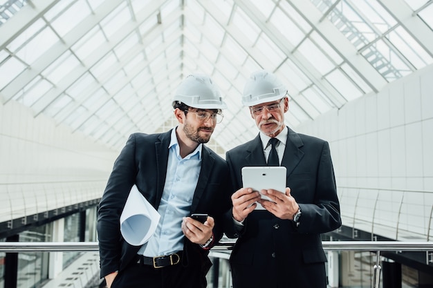 Бесплатное фото Два успешных предпринимателя в шлеме с проектом и ноутбуком новостройки остаются возле стеклянной крыши