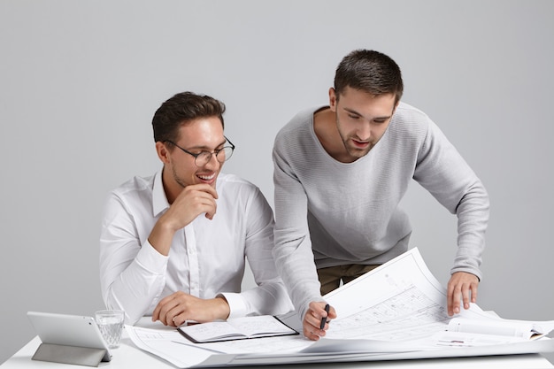 Two successful professional male constructors studying blueprints at spacious light office