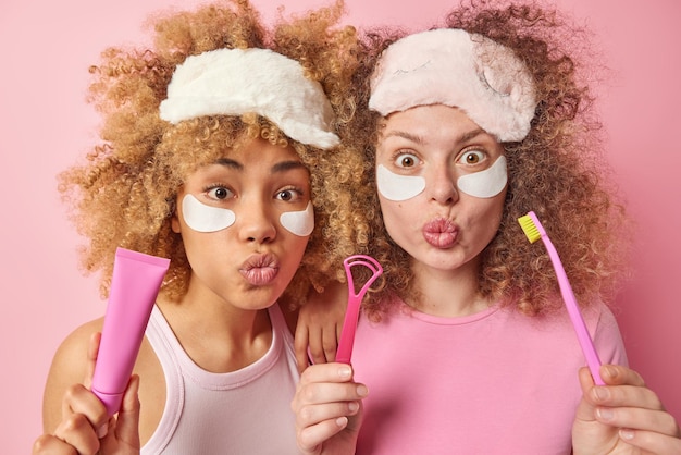 Due sorelle femmine sorprese con i capelli ricci indossano maschere per dormire applicano cerotti di bellezza sotto gli occhi andando a pulire i denti e la lingua con la spazzola stanno vicini l'uno all'altro isolati su sfondo rosa