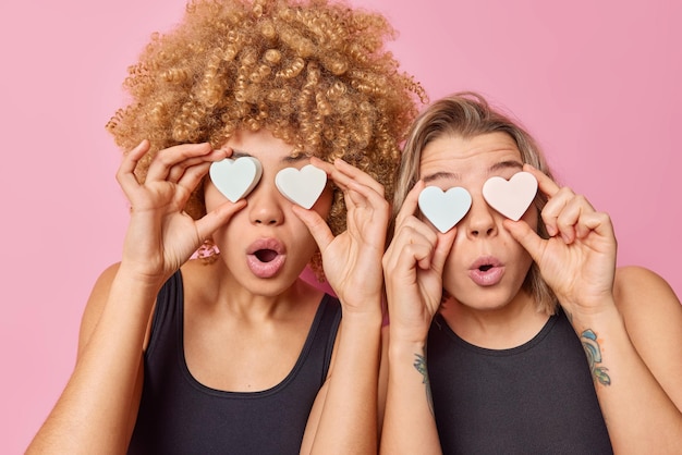 Free photo two surprised women cover eyes with heart shaped soap keep mouthes widely opened from wonder take care of skin and personal hygiene dressed in black t shirts stand next to each other indoor