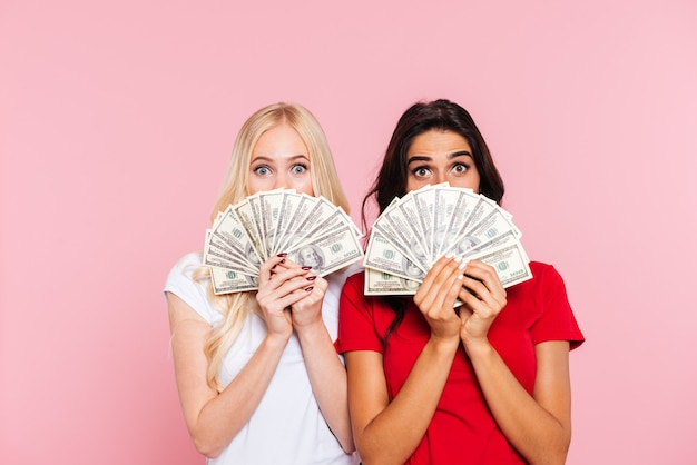 Two surprised women hiding behind the money and looking at the camera over pink 