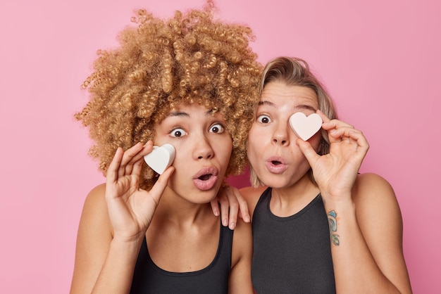 Free photo two surprised women hold heart shaped soaps take care of skin and complexion look with widely opened eyes and mouth dressed in casual black t shirts isolated over pink background wellness concet