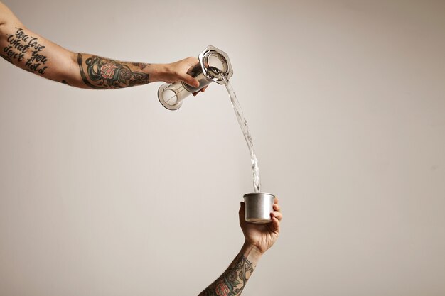 Two tattooed man's hands pouring water from a clear plastic aeropress into a small steel travel cup on white Alternative coffee brewing commercial