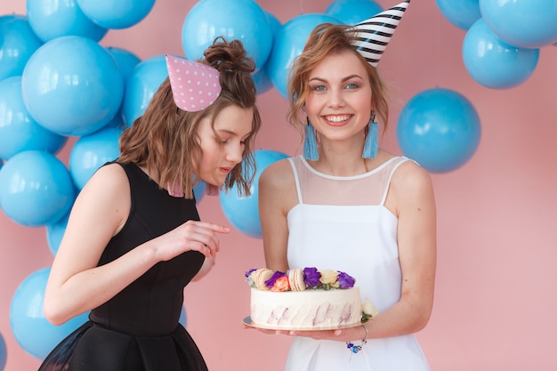 Two teenage girls in party hat holding cake. Isolated on pink background and blue balloons