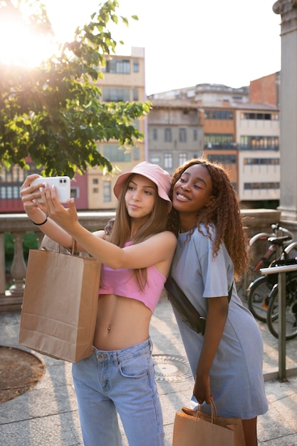 Две девочки-подростки делают селфи после похода по магазинам, держа в руках сумки