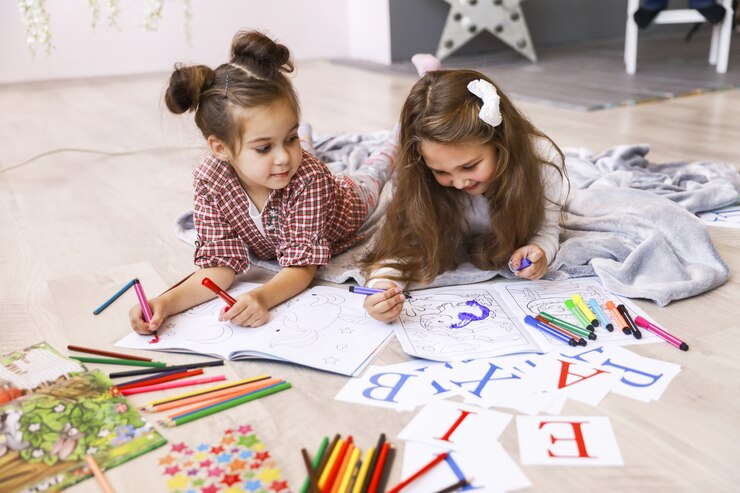 Free Photo two tiny happy girls which are drawing in the coloring book laying on the floor on the blanket and learning letters
