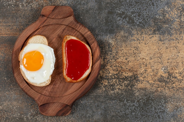 Two toast bread with fried egg and jam on wooden plate.