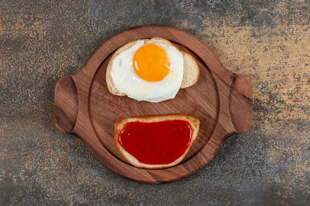 Two toast bread with fried egg and jam on wooden plate.