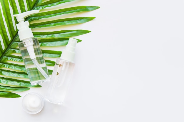 Two transparent cosmetic bottle with spray head and moisturizer on green leaf against whit e background