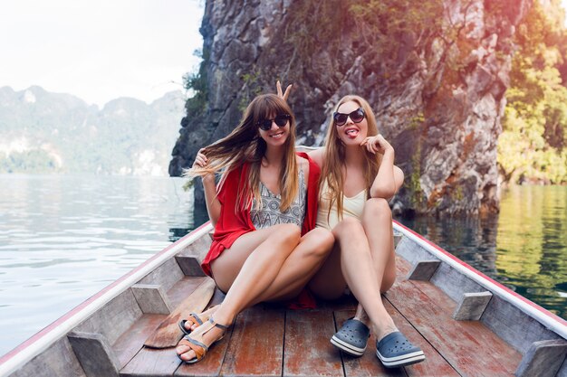 Two traveling women, best friends exploring wild nature of Khao Sok national park. Sitting in wood long tail boat on tropical limestone cliffs. Lifestyle image. Island lagoon.