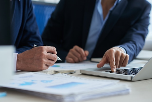 Free photo two unrecognizable male colleagues using laptop and business charts lying on desk