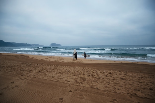 Two unrecognizable surfing girls with their longboards stay on ocean shore and watching waves in early morning, wearing full wetsuits and ready to surf