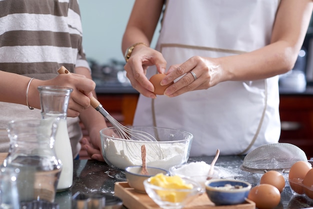 Two unrecognizable women holding whisk and breaking egg into mixing bowl