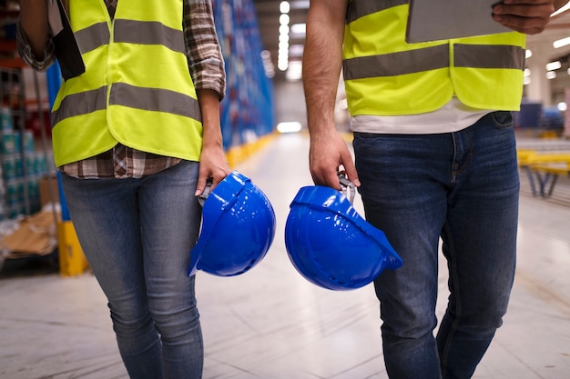 Two unrecognizable workers in reflective suit walking through warehouse and holding blue protective hardhats