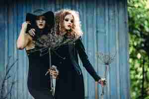 Free photo two vintage women as witches, posing in front of an abandoned house on the eve of halloween