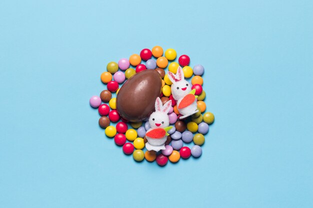 Две белые кролики и шоколадное пасхальное яйцо на жемчужинах конфет на синем фоне