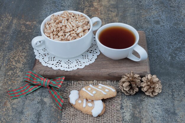 Two white cups with Christmas cookies and two pinecones on wooden board. 