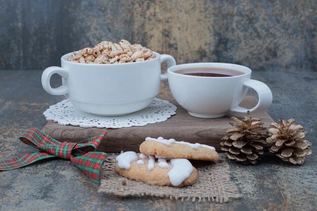 Two white cups with Christmas cookies and two pinecones on wooden board.