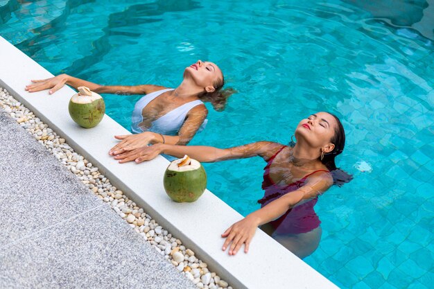 Two woman friends asian and caucasian with makeup in swimming pool at villa