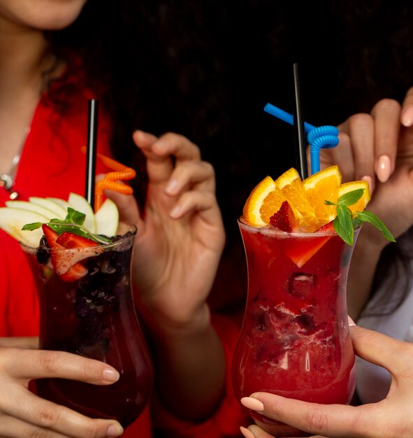 two woman holding glasses of fruit cocktails with orange, strawberry and apple, strawberry