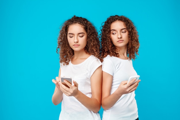 Two womans twins looking at phones over blue.