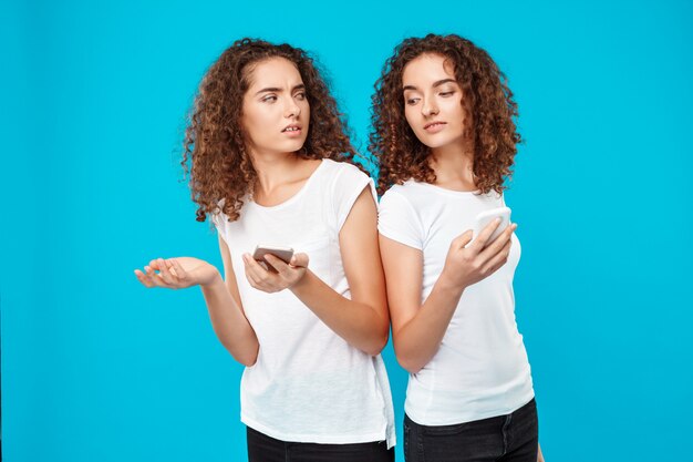 Two womans twins looking at phones over blue.