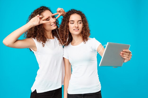 Two womans twins making selfie on tablet over blue.