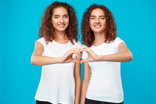 Two womans twins showing heart with hands over blue.