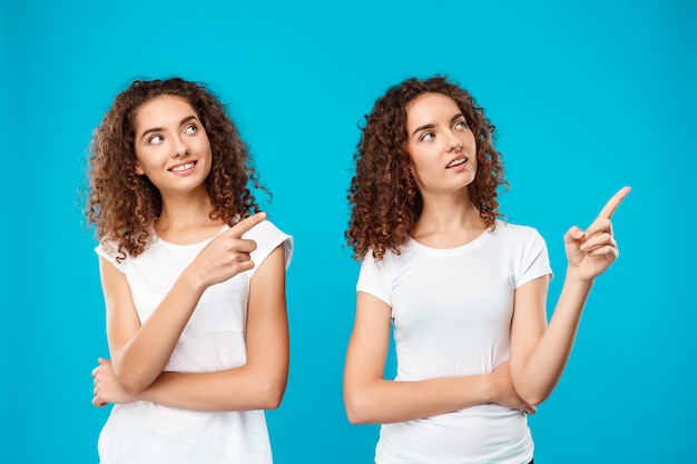 Two womans twins smiling, pointing fingers away over blue.