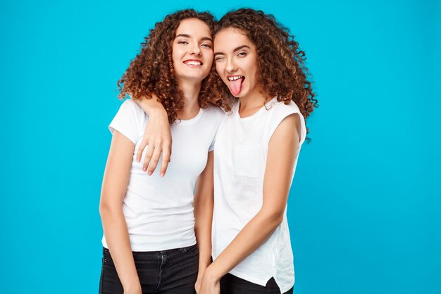 Две женщины близнецы улыбаясь, показывая язык над синим.