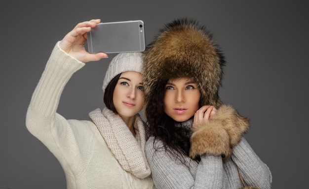 Two womans in winter hats taking pictures on smartphone. Isolated on grey background.