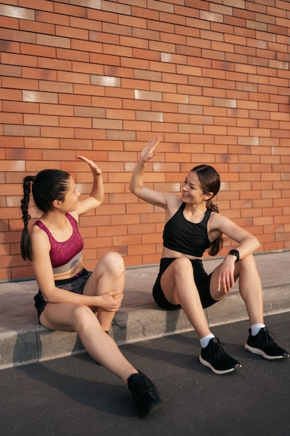 Two women after urban workout giving hi five for excellent results. Girls preparing for running and sitting in the street.