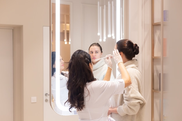 Two women, a beautician doctor and a client, stand at the mirror, at a consultation, discussing the upcoming procedures. Beautician talks about sculpting the face