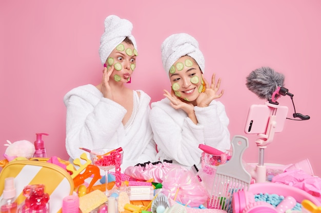 Two women bloggers show how to make natural face mask apply cucumber slices on face and record vlog video on smartphone wear white soft bathrobes and towels over head isolated over pink wall