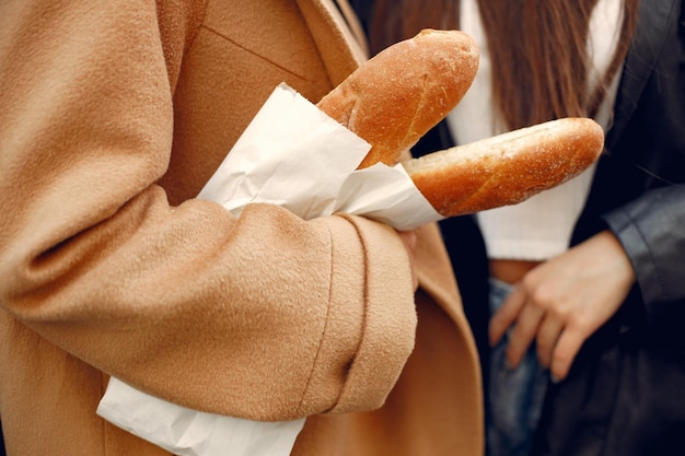 Two women in coats on a street. Cropped photo of person holding french baguettes in kraft paper bag in hands. Women wearing brown coat and black leather coat.