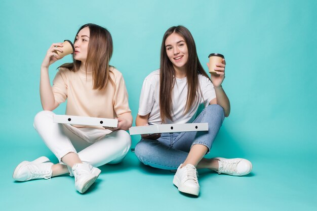 Two women friends sitting on the floor drink coffee to go and eat pizza isolated on turquoise wall