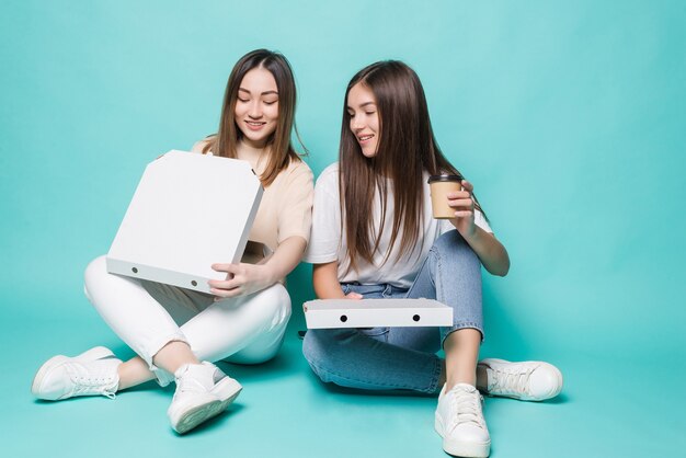 Two women friends sitting on the floor drink coffee to go and eat pizza isolated on turquoise wall