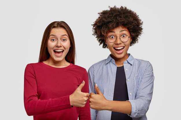 Two women give thumbs to each other, show their agreement, smile gladfully, stand shoulder to shoulder over white wall, gesture indoor. Interracial friends being glad, give okay sign, feel overjoyed