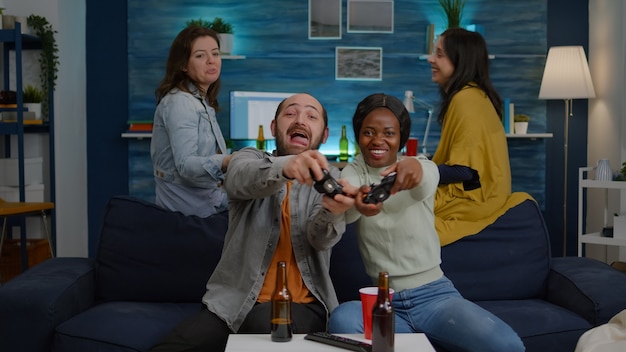 Two women losing online videogames using joystick playing for gaming competition. Multi-ethnic friends drinking beer, socialising, having fun together sitting on couch late at night