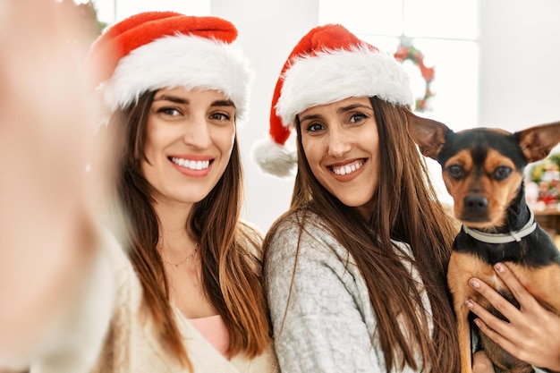 두 명의 여성이 집에서 크리스마스 장식 옆에 서 있는 개를 안고 있는 카메라 옆에서 셀카를 만듭니다.