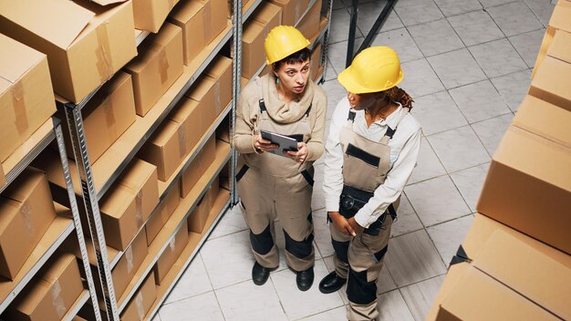 Two women in overalls working with industrial goods, planning shipment of products with digital tablet. Team of employees doing inventory in storage room, manufacturing products.