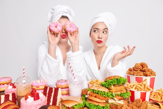 Two women pose near table full of delicious appetizing snacks prefer cheat meal istead of healthy one.