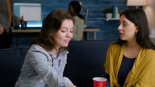 Two women relaxing on sofa drinking beer while socializing during home party