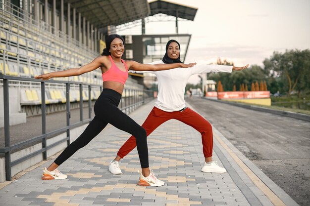 Two women on a stadium doing stretching. Muslim woman in hijab wearing white shirt and red trousers