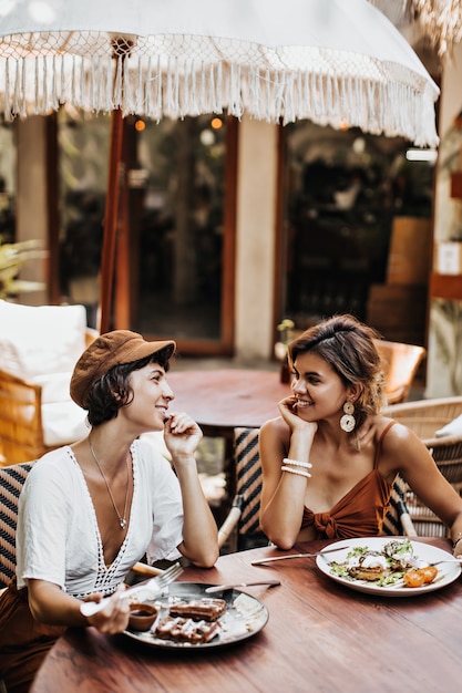 Two women in summer stylish outfits talking and eating delicious food in street cafe