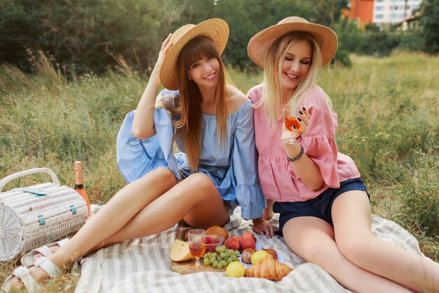Two wonderful girls in straw hat spending holidays in countryside, drinking sparkling wine.