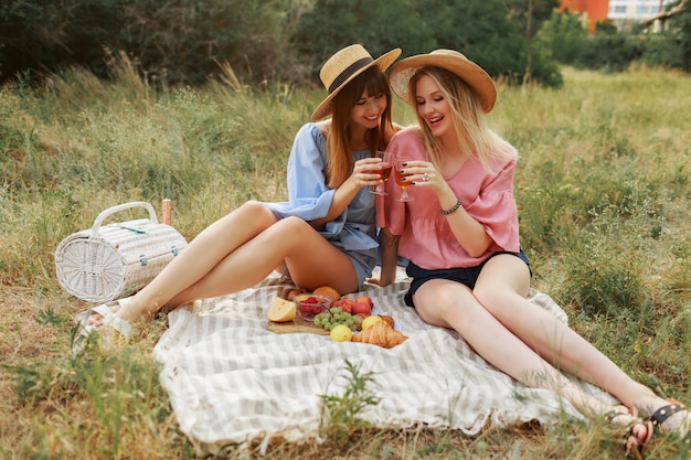 Two wonderful girls in straw hat spending holidays in countryside, drinking sparkling wine.